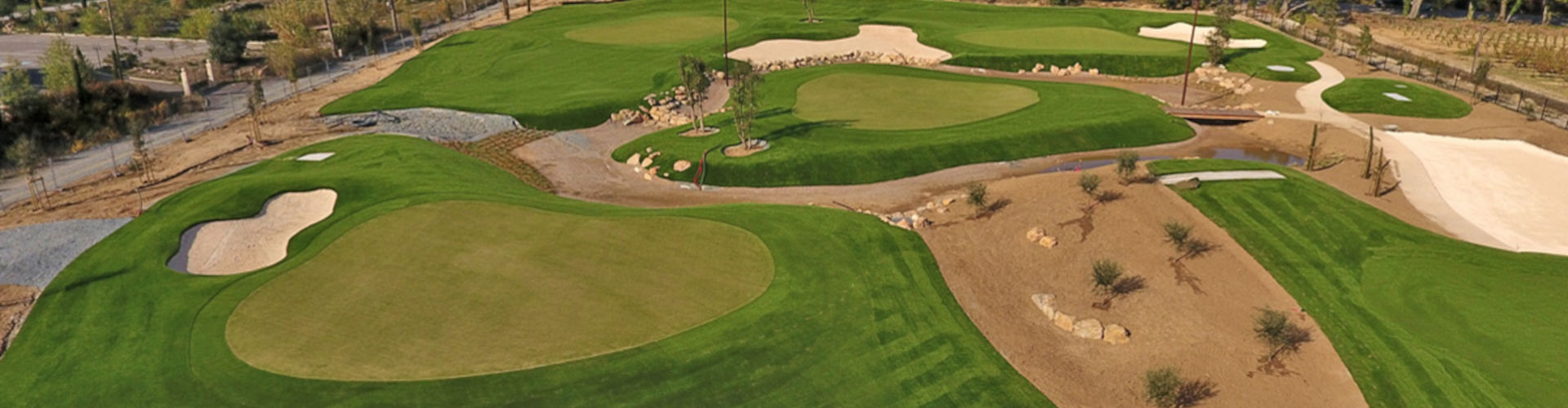 Aerial view of a commercial putting green turf installation with multiple holes, sand bunkers, rock features, and vineyard and mountain views in the background
